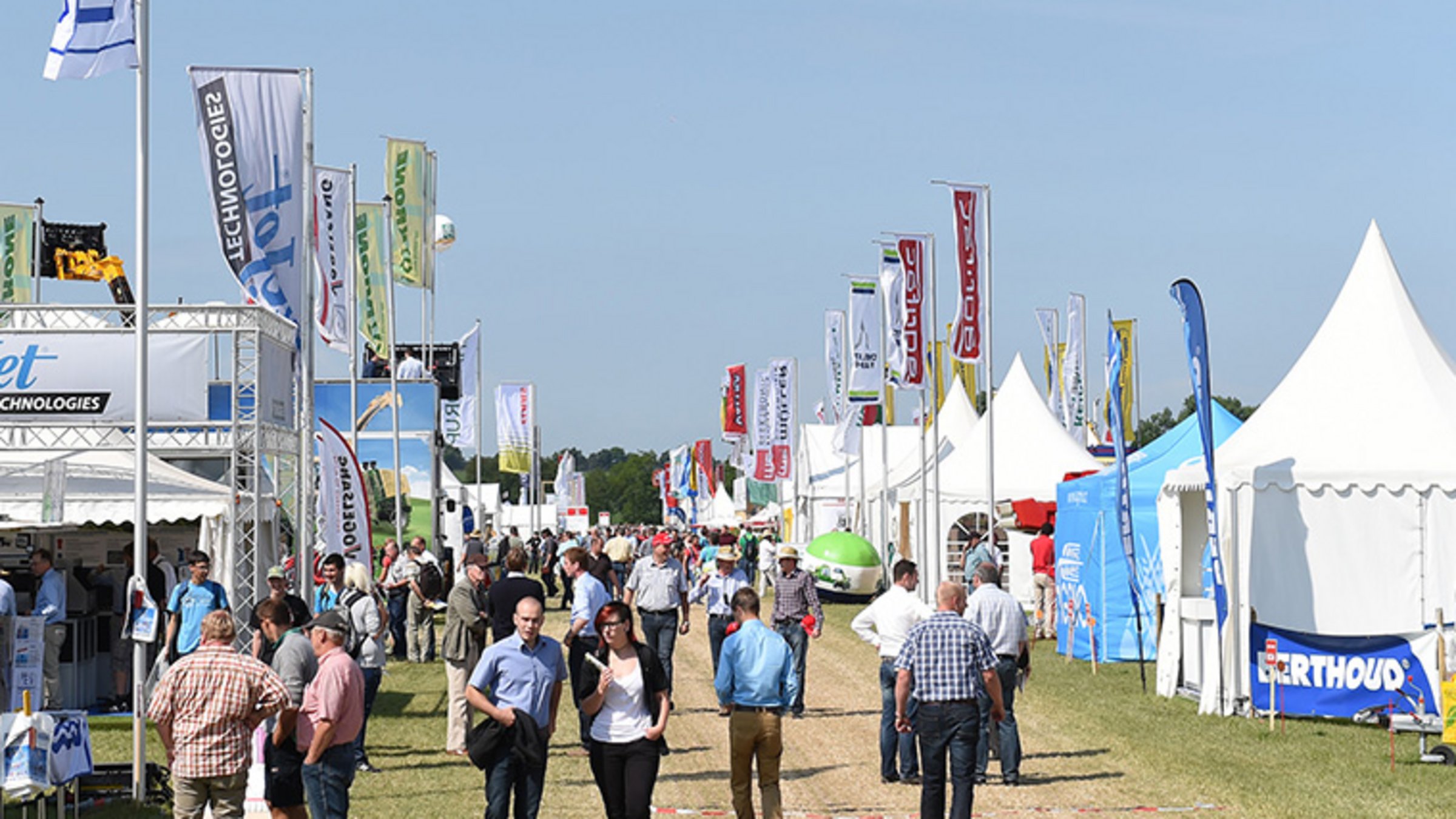 Agricultural open-air fair with visitors, tents, and exhibitor banners on a sunny day.